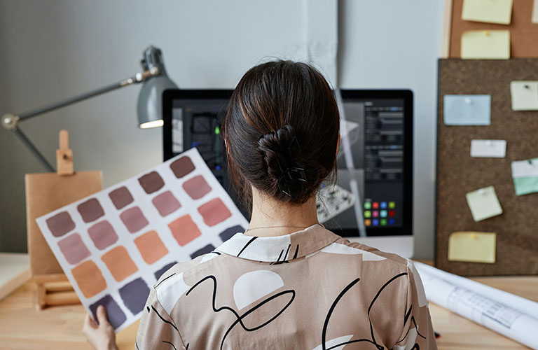 A woman holds up colour swatches while reviewing floor plans on a computer.