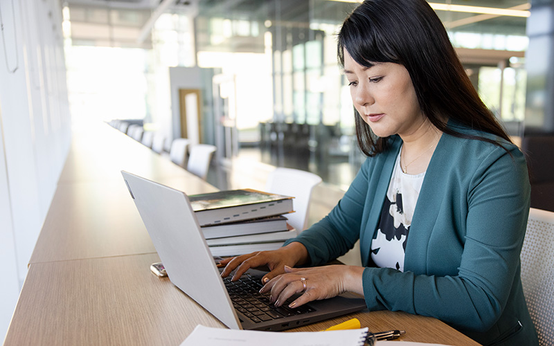 A woman works on a laptop at a tall long desk in a glass and metal building.