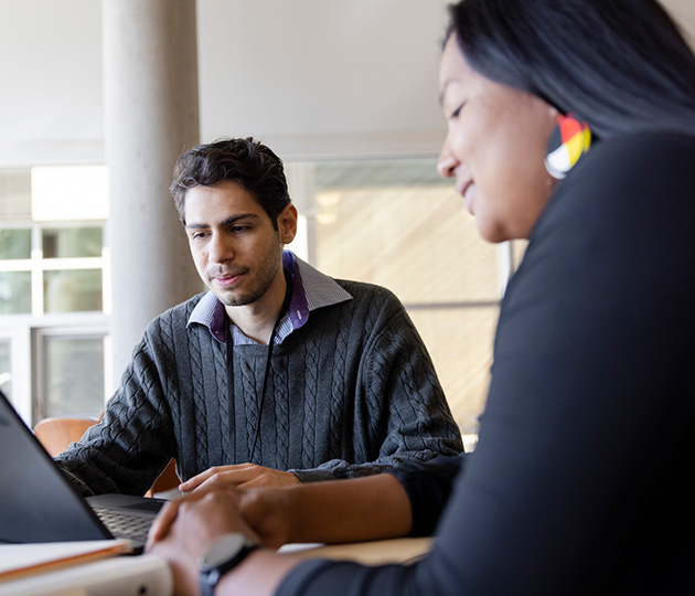 A man in professional clothing looking at a laptop beside a woman wearing medicine wheel earrings.