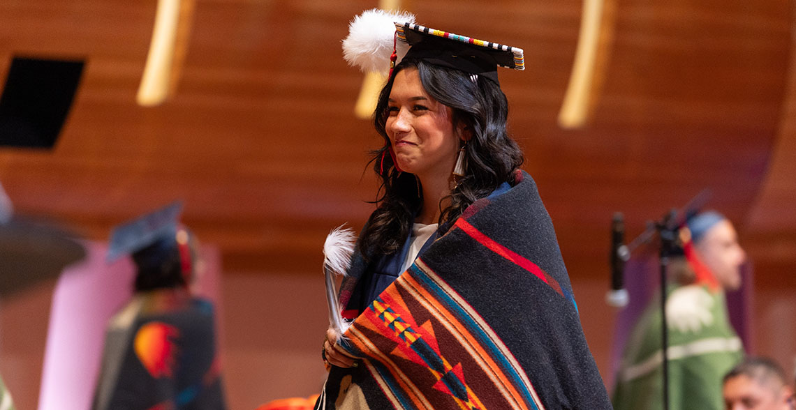 A student in grad cap decorated with beads and a feather, a graduation gown, wrapped in a cerimonial blanket at Indigenous Honouring Ceremonies.