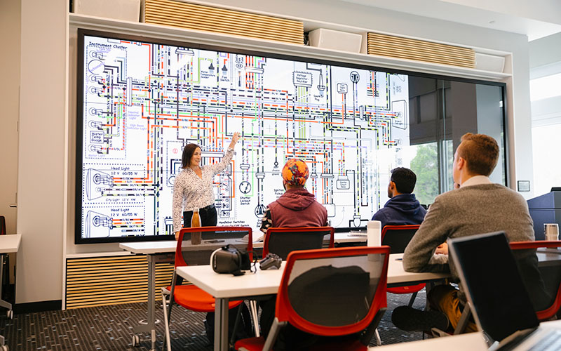 A professor teaching in front of a wall of screens in a classroom.