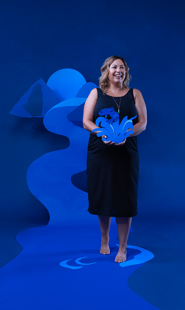 Photo of Jenny Philbrick in front of a blue backdrop. A mountain scene made of cut out pieces of blue paper is arranged behind her. She is holding a blue paper cut out of a fish leaping from a splash of water.