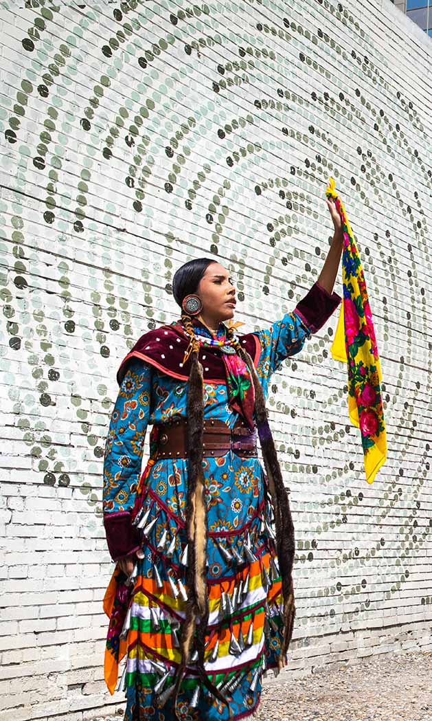 Sofia Baptiste, wearing a jingle dress, hand raised in the hair in front of a mural of many dots arranged in a circle.
