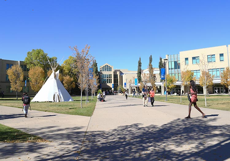 Photo of East Gate in fall. Students walk around. A teepee is seen on the lawn.