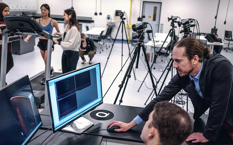 Researchers of the Foot-Ankle Stability Lab (FASt Lab) reviewing data on a laptop as a participant walking on a treadmill while being measured by many cameras.