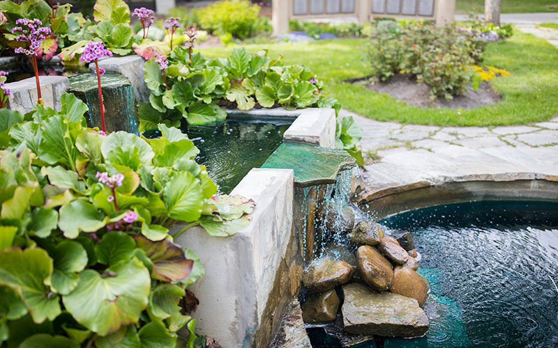 A waterfall pouring over landscaped rocks that are surrounded by flowers and other plants.