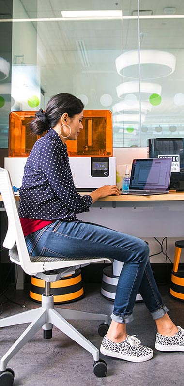 Female student at a desk, working on a laptop with a 3D printer in the background.