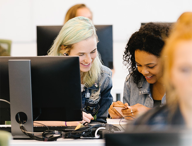 In a bright computer lab, two students are smiling while comparing class notes.