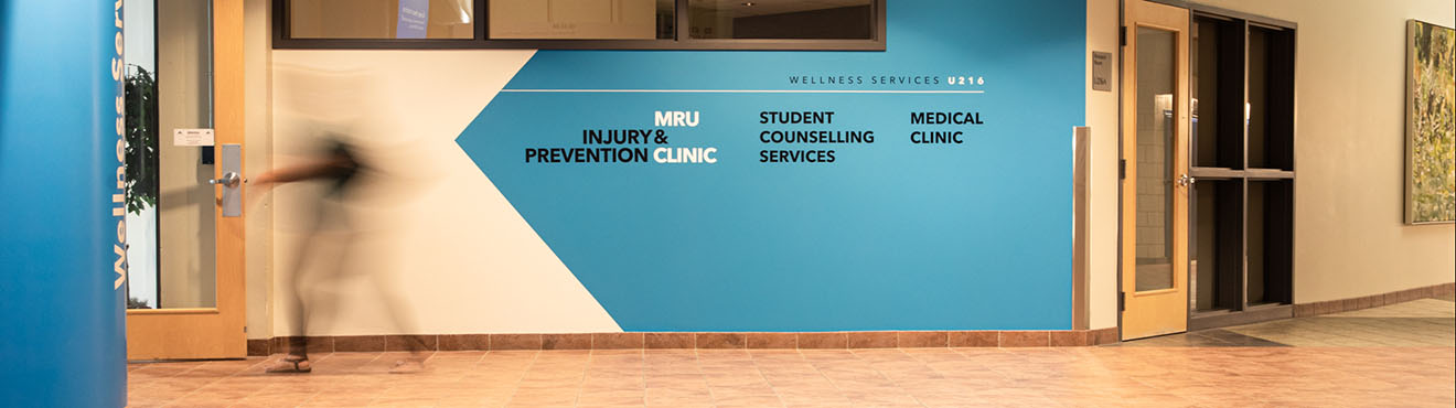 Photo of the entrance to the MRU Injury & Prevention Clinic and Wellness Services. The department names are on the wall in vinyl and a person in motion is walking by.