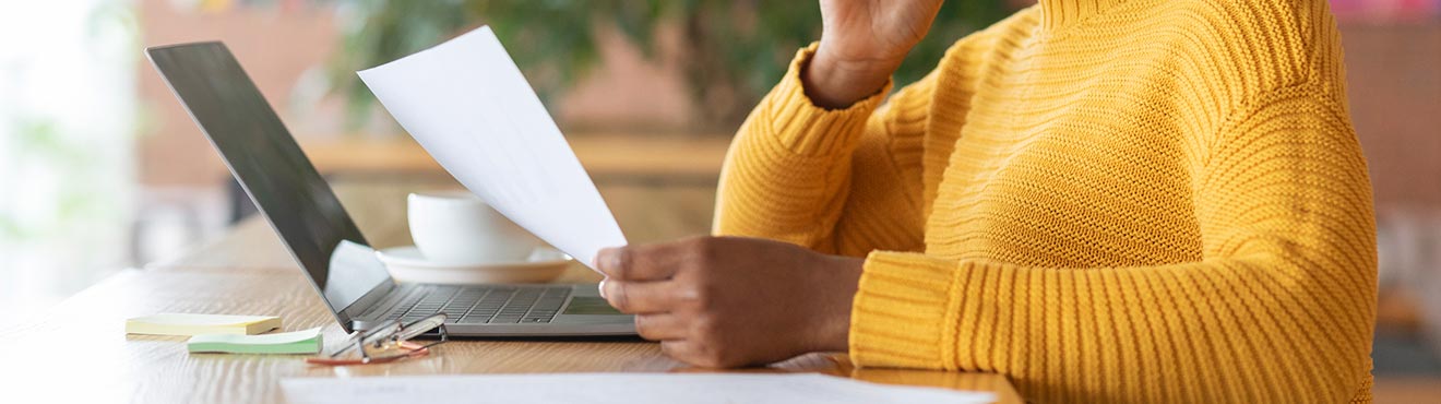 A person reviewing a piece of paper on a desk covered in office goods and a laptop.