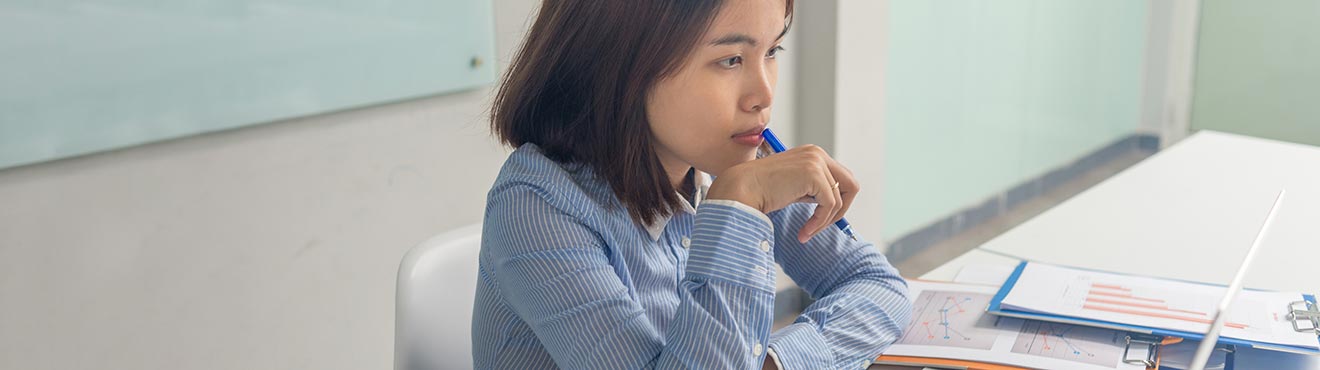 A woman in business attire thinking while sitting at a desk surrounded by charts and graphics.