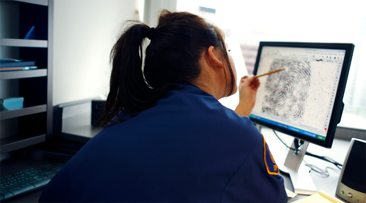 A woman sits at a computer, rexamining an enlarged finger print on the screen.