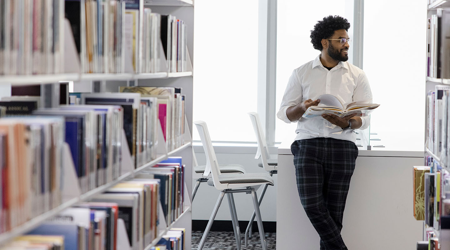 A man in a white button-up shirt stands leaning on a table, holding an open book in between the stacks of the Riddell Library and Learning Centre.