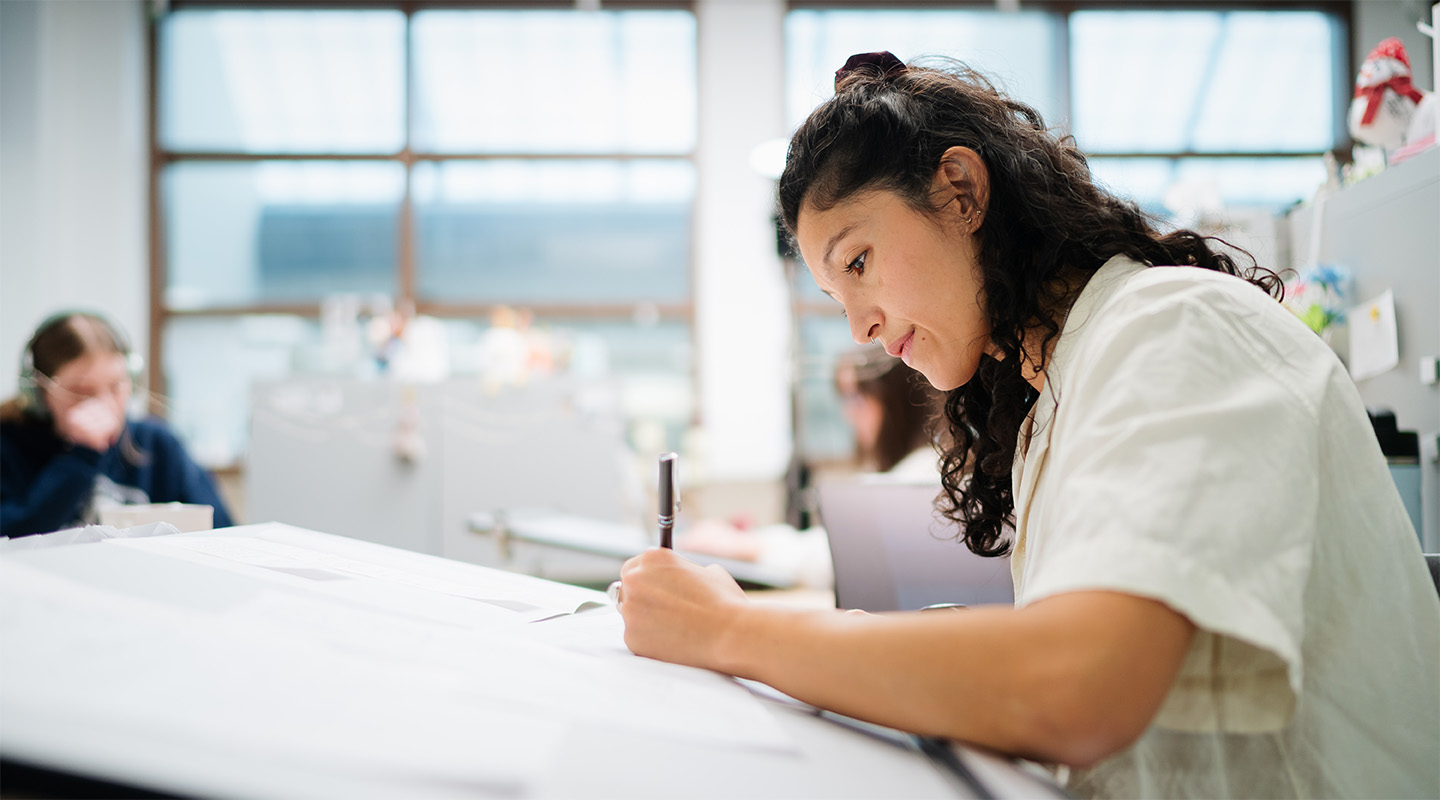 A woman with curly hair sits at a drawing desk, focused on her project.