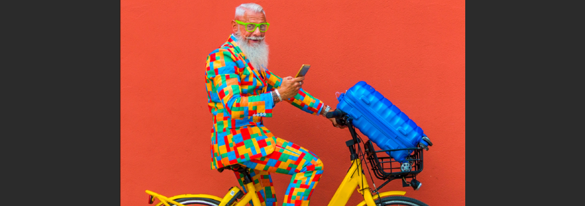 older adult male wearing a colourful suit is riding a yellow bicycle with bright blue luggage in front-of-bike basket
