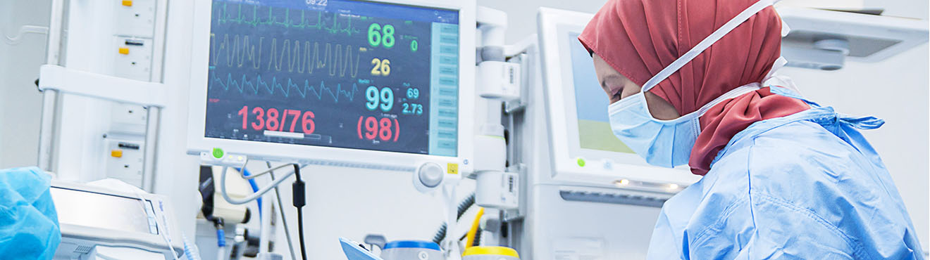 A nurse wearing a hijab reviews a chart in a patient room full of various medical equipment.