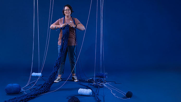 Photo of Rachael Edwards in front of a blue backdrop knitting a large blue scarf.