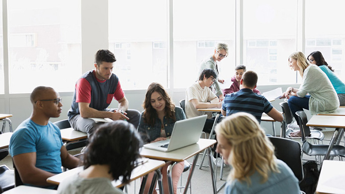 Young woman using laptop with students watching and smiling.