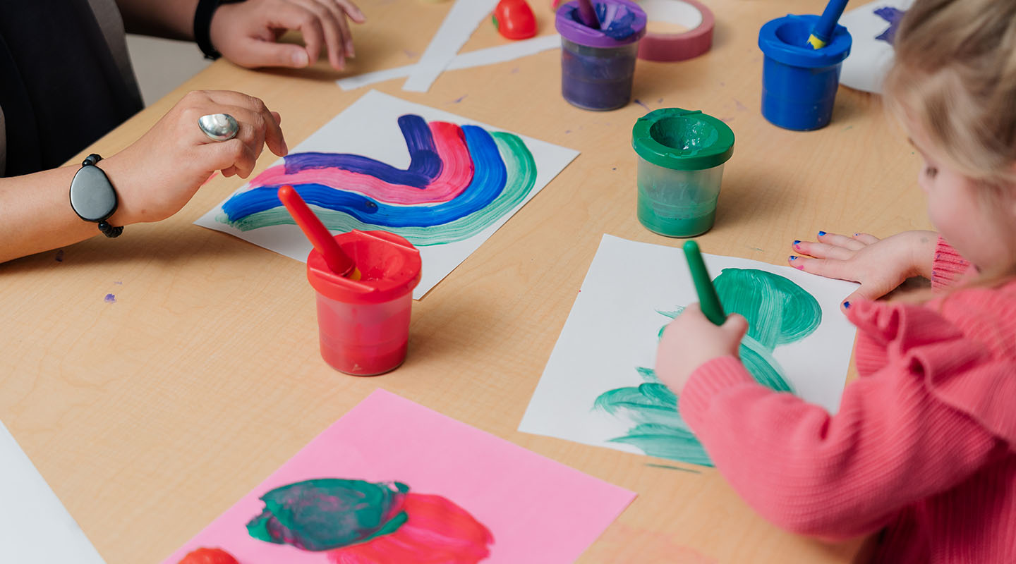 A young child sits at a desk colouring while a child care professional sits across from her.