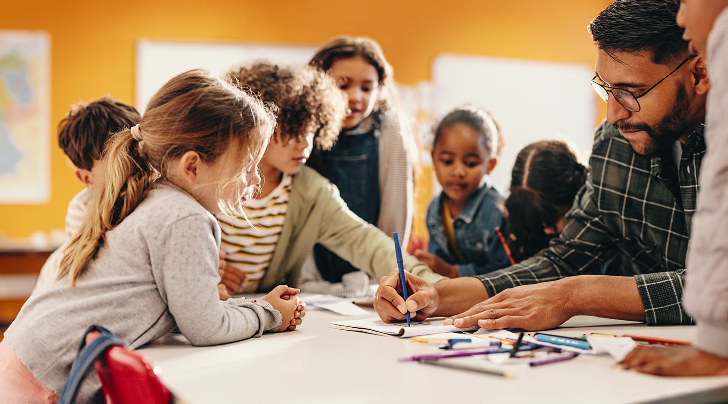 A group of elementary aged students gather around their teacher at a low table to see a drawing the teacher has made.