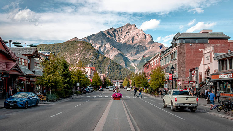 Photo of the main street in Banff, Alberta. A road with people walking along shops with a mountain in the background.