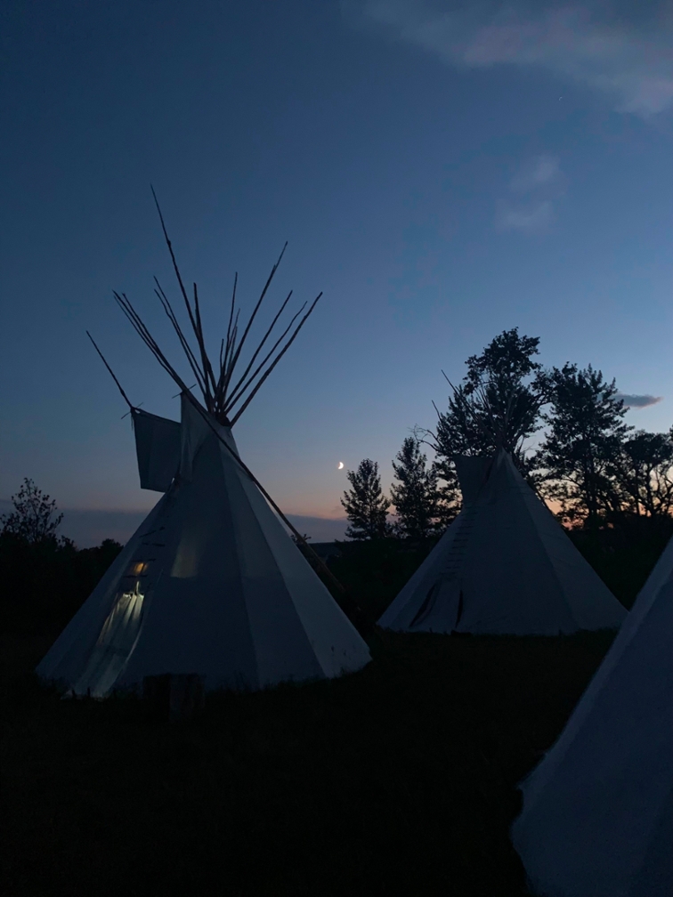 A few tipis right after sunset. There is a cresent moon in the sky in the background.
