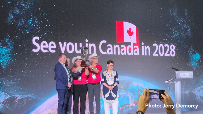 Five people stand on a stage in front of a screen that reads 'See you in Canada in 2028.' The middle two are holding up a trophy.