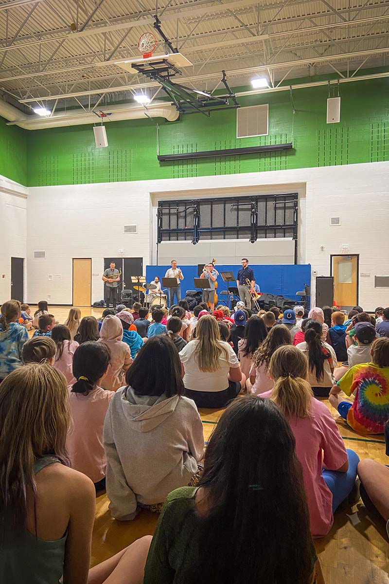 A gym full of primary school students sitting on the floor while Conservatory instructors perform at an Outreach Clinic.