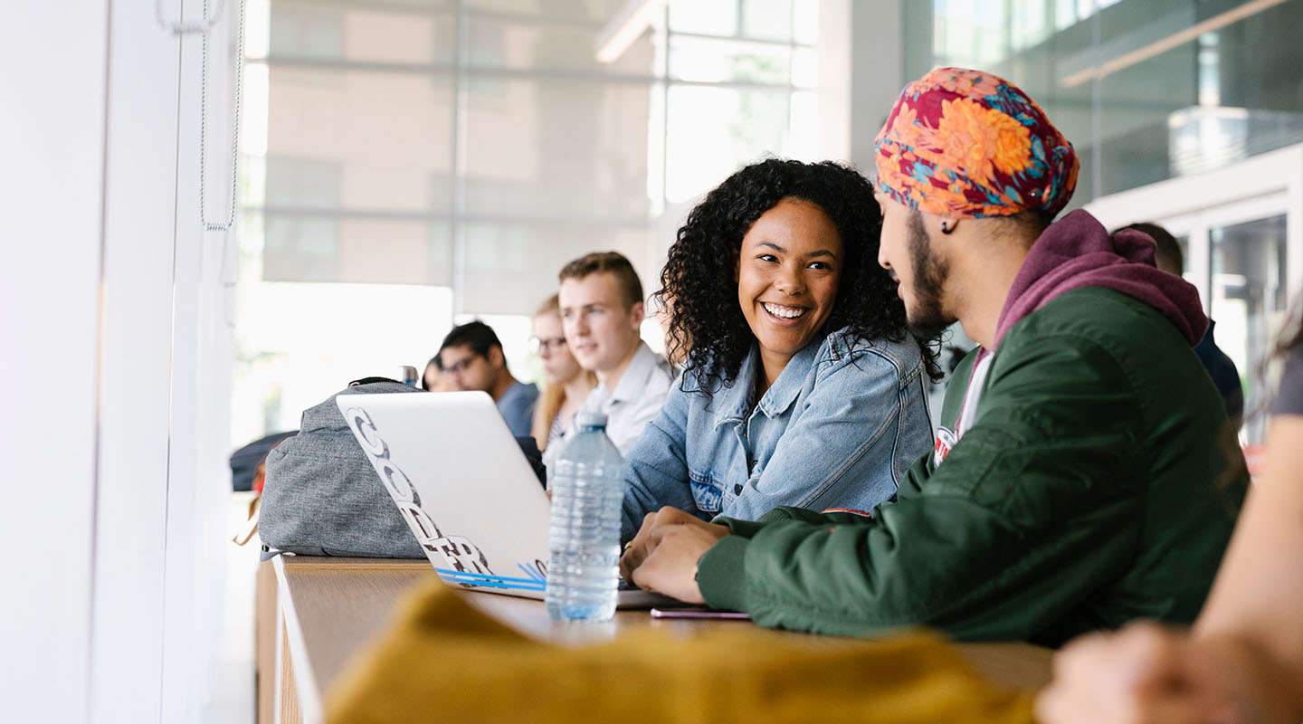A young curly haired woman and a young man in a brightly coloured turban having a conversation in the Riddell Library and Learning Centre.