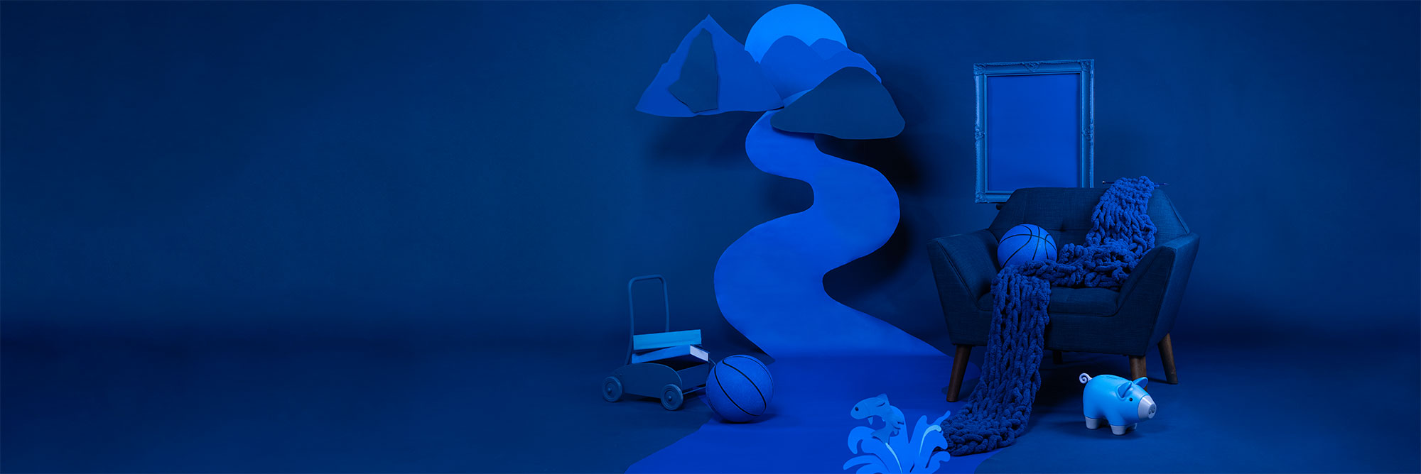 A blue background with a chair, backetball, picture frame, children's toys and scarf: all blue. A mountain scene with a river is cut out of different shades of blue paper and hung in the background.