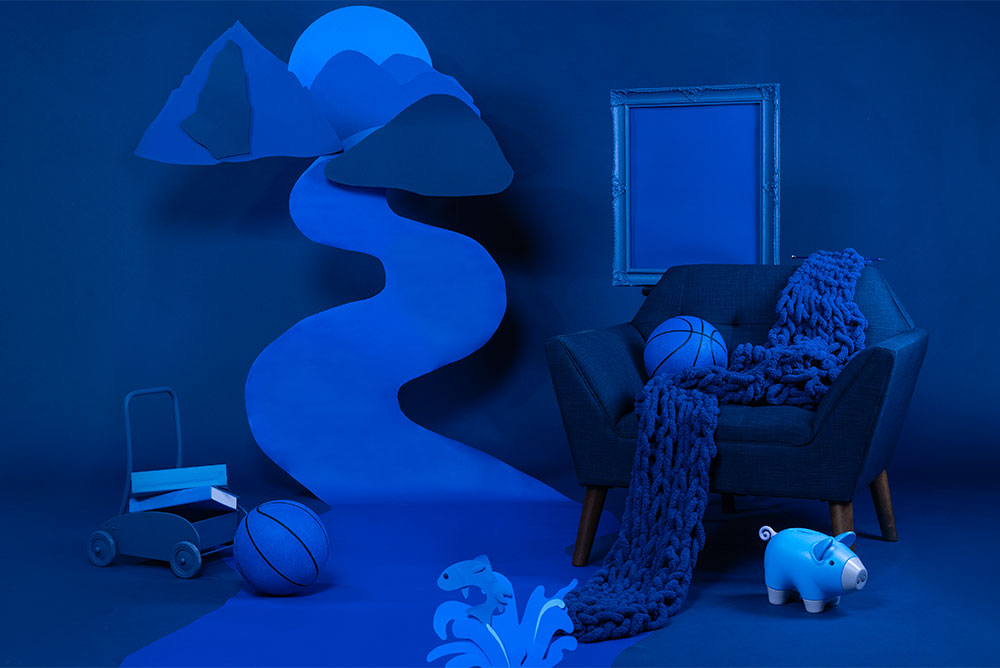 A blue background with a chair, backetball, picture frame, children's toys and scarf: all blue. A mountain scene with a river is cut out of different shades of blue paper and hung in the background.