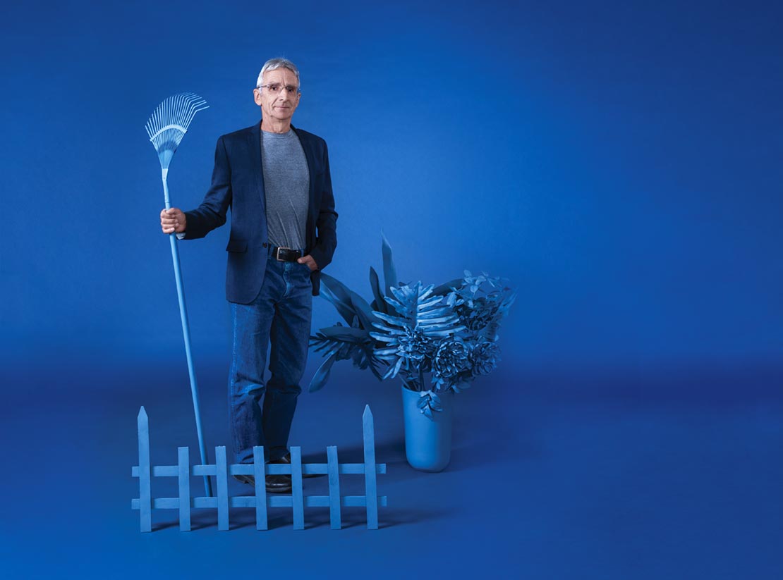 Walter Hossli on a blue backdrop holding a blue painted rake beside a blue painted plant and picket fence.