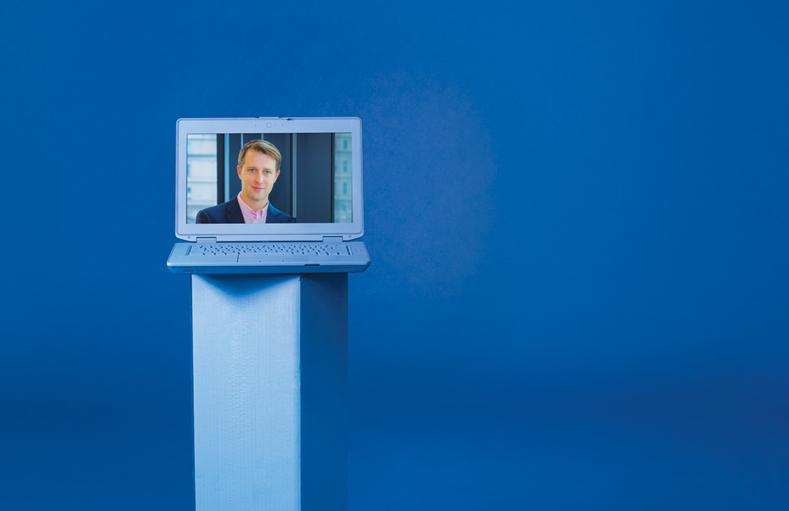 Photo of Geoff Schoenberg on the screen of a blue painted laptop placed on a blue painted pedestal on a blue backdrop.