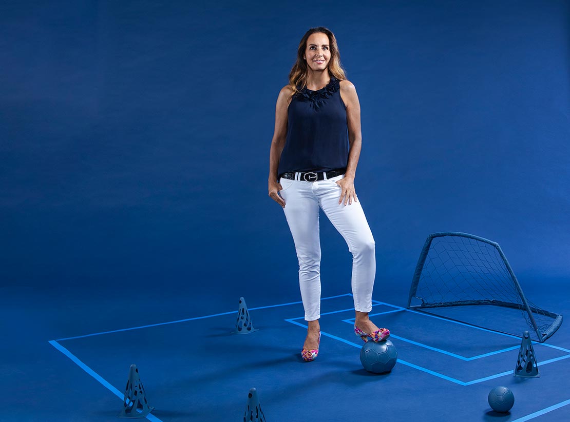 Photo of Jacquie Hertlein on a uniformly blue backdrop. There are a variety of sports objects painted blue around as she props a foot on a blue painted soccer ball.