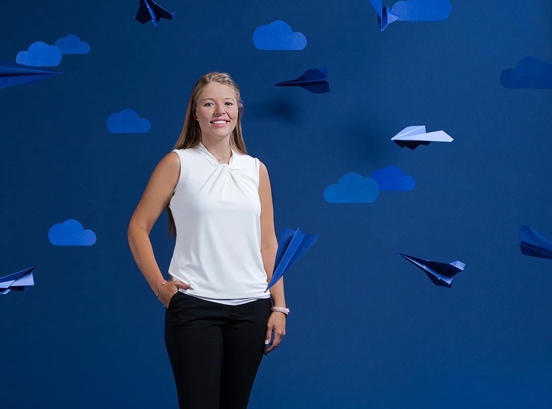 Photo of Sophia Wells on a uniformly blue backdrop. She is surrounded by blue paper airplanes and clouds cut out from blue construction paper.