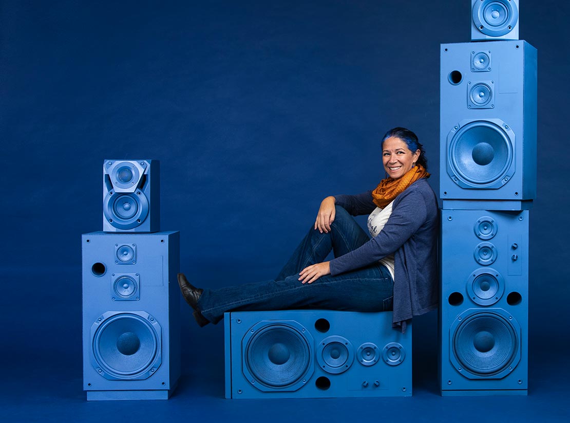 Photo of Heather Graham on a uniformly blue backdrop. She is sitting on a blue painted speaker and is leaning against a larger stack of blue painted speakers.