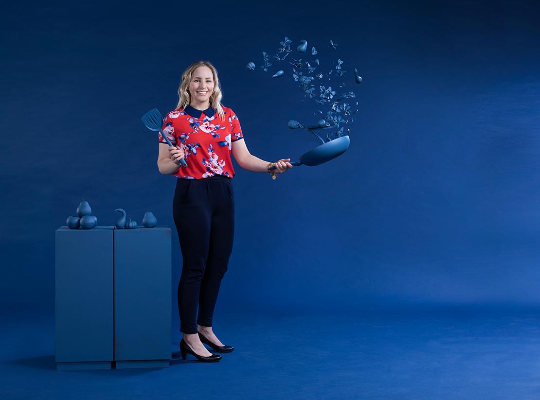 Photo of Robyn Madden on a uniformly blue backdrop. She is tossing blue painted food out of a blue wok into the air and holding a blue spatula. In the background blue painted food rests on a blue table.