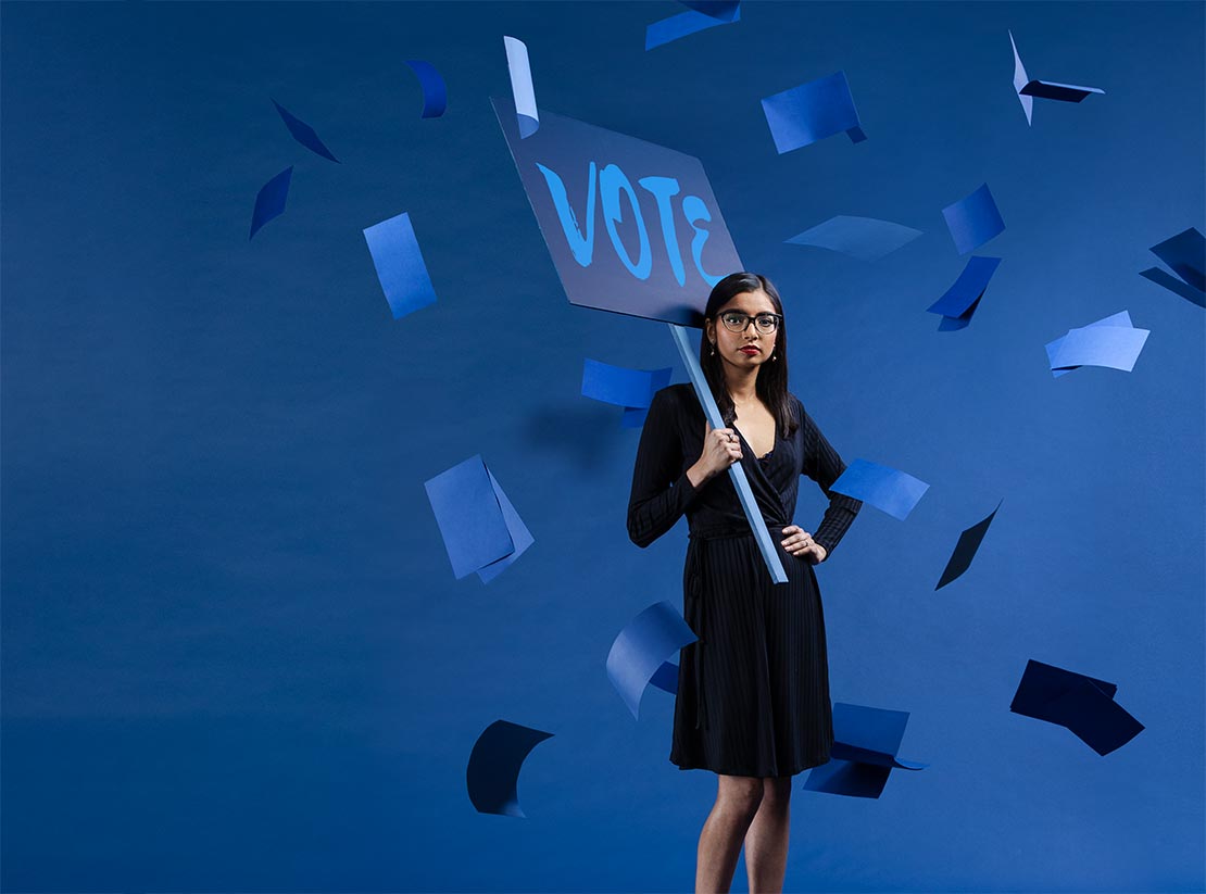 Photo of Shifrah Gadamsetti on a uniformly blue backdrop. She holds a blue picket sign with the word vote writtten on it. Blue construction paper floats down around her.