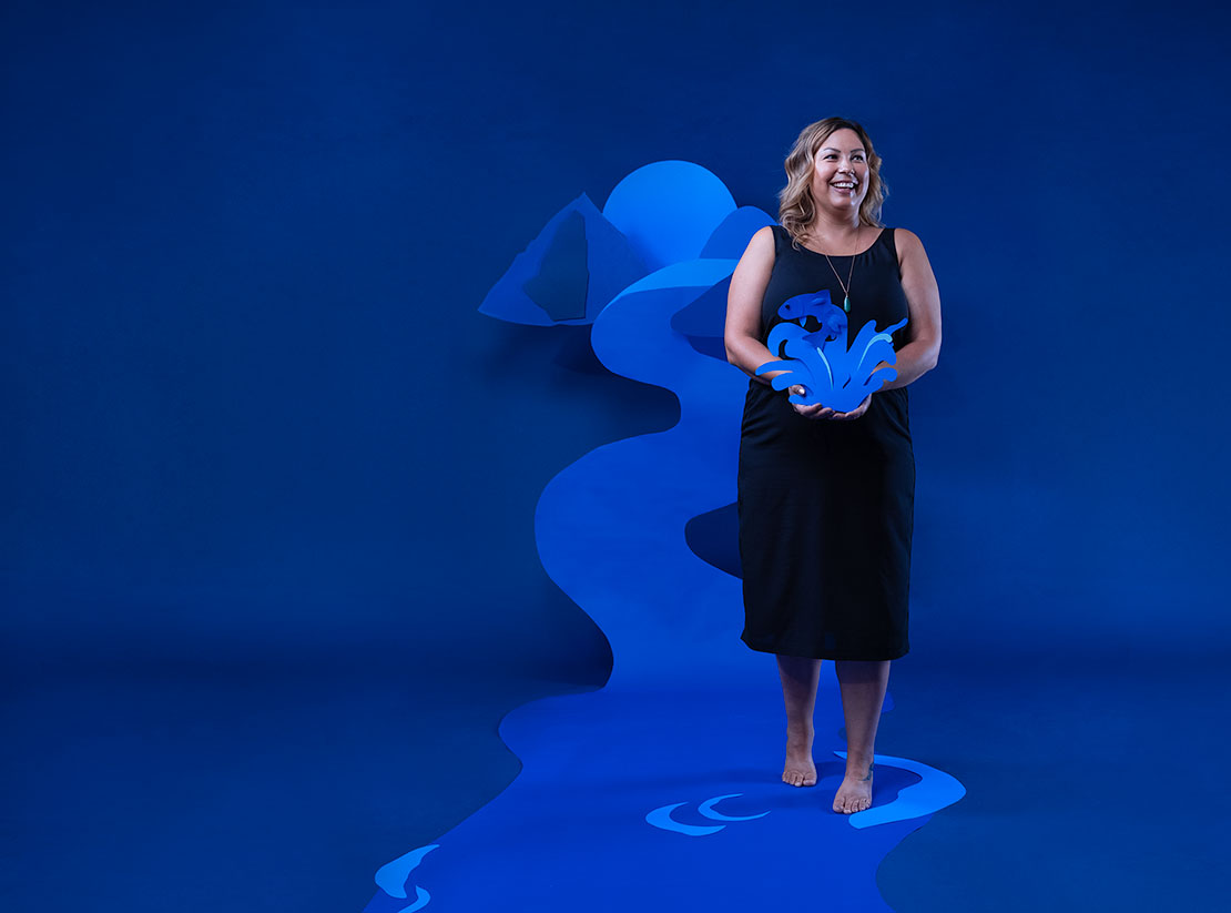 Photo of Jenny Philbrick in front of a blue backdrop. A mountain scene made of cut out pieces of blue paper is arranged behind her. She is holding a blue paper cut out of a fish leaping from a splash of water.