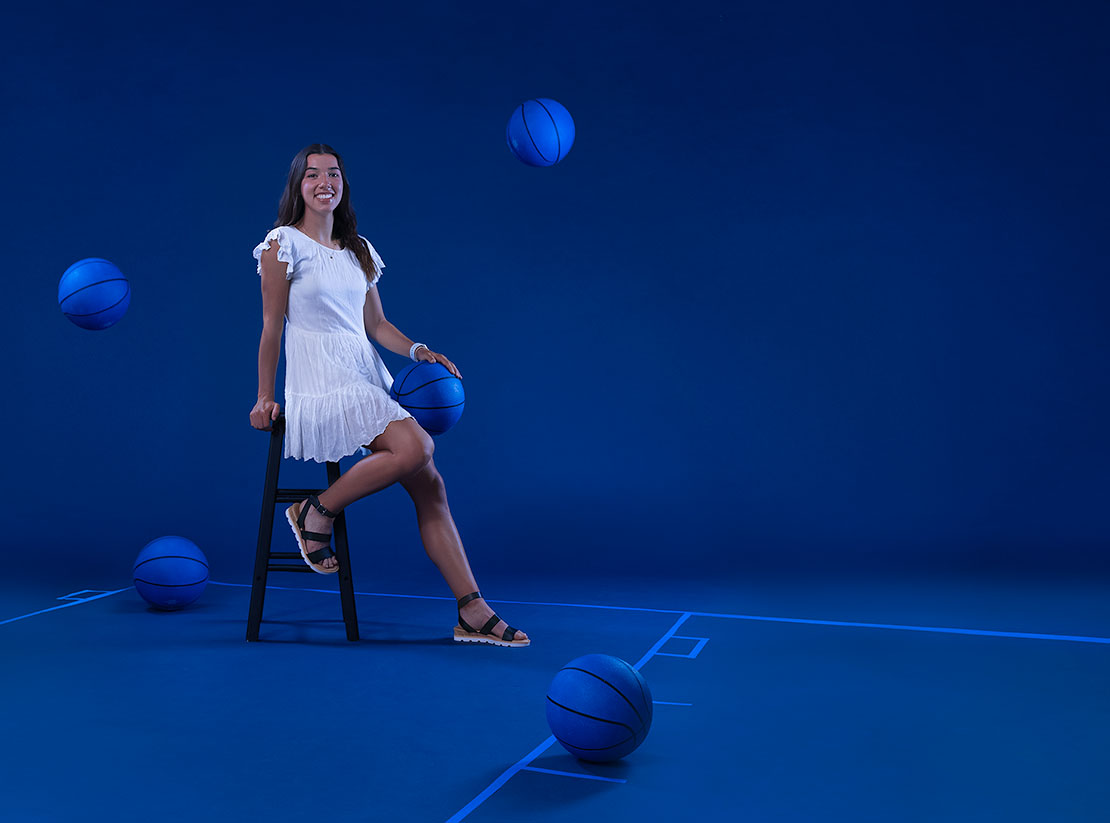 Photo of Marnie Garner in front of a blue backdrop. She sits on a stool holding a blue painted basketball. Basketball court lines are taped out on the floor and other blue basketballs bounce around.