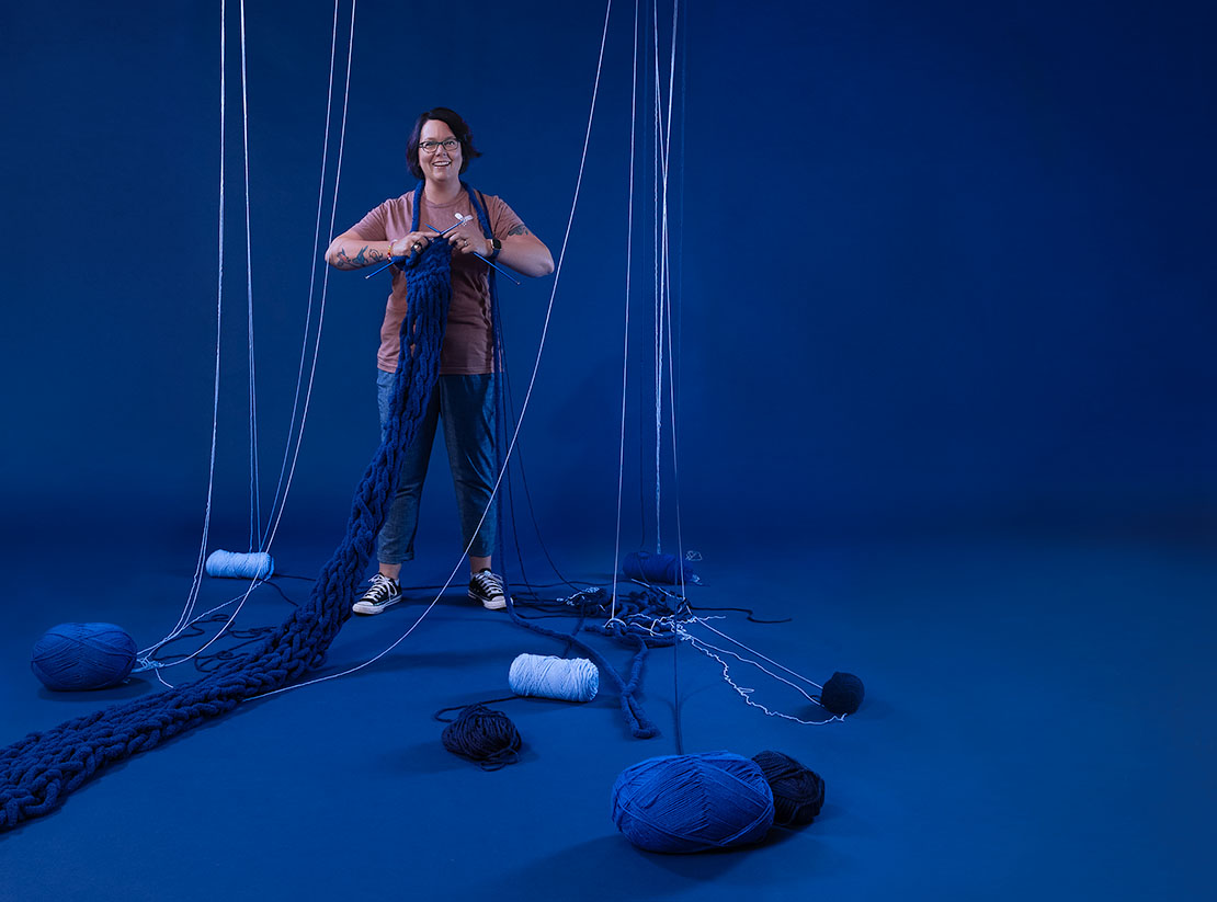 Photo of Rachael Edwards in front of a blue backdrop. She is holding oversized, blue knitting needles and appears to be knitting a large blue scarf. Blue yarn is strewn about.
