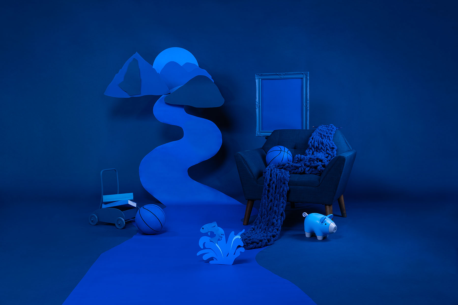 A blue background with a blue chair with a blue backetball on the seat and blue yarn scarf drapped over it. A mountain scene with a river is cut out of different shades of blue paper and hung in the background. There is a blue frame hung and blue children's toys scattered about.