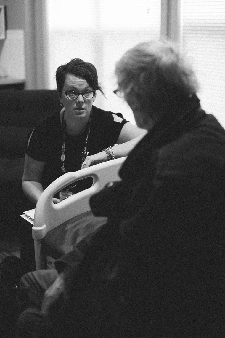 Black and white photo of Rachel Edwards leaning in and conversing with Jerome Domanski, who sits on a medical bed