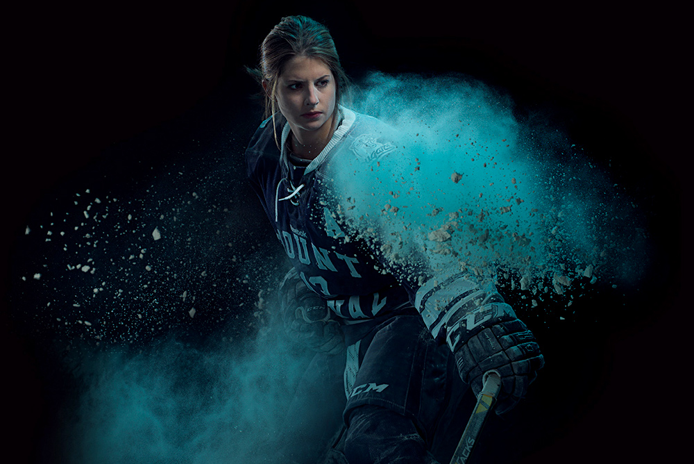 A member of the Women's Cougar's Hockey team in goalie gear takes a defensive stance in dramatic lighting and blue powder.