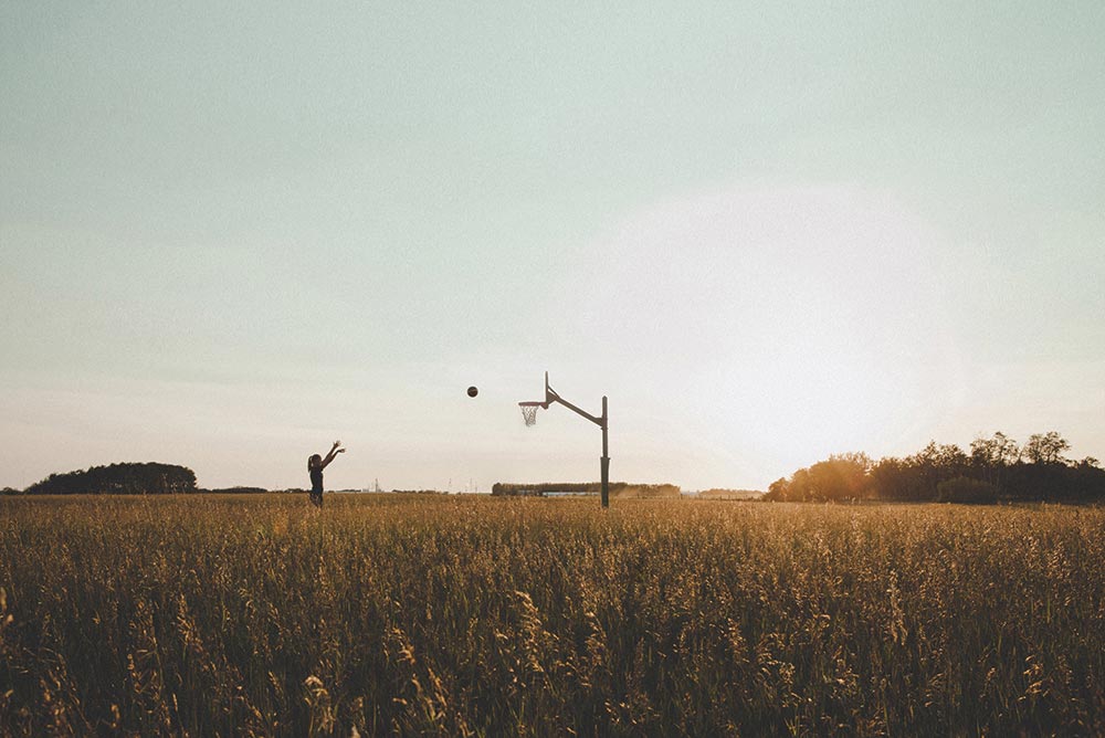 A young woman stands in a rural field and shoots a basketball at a hoop