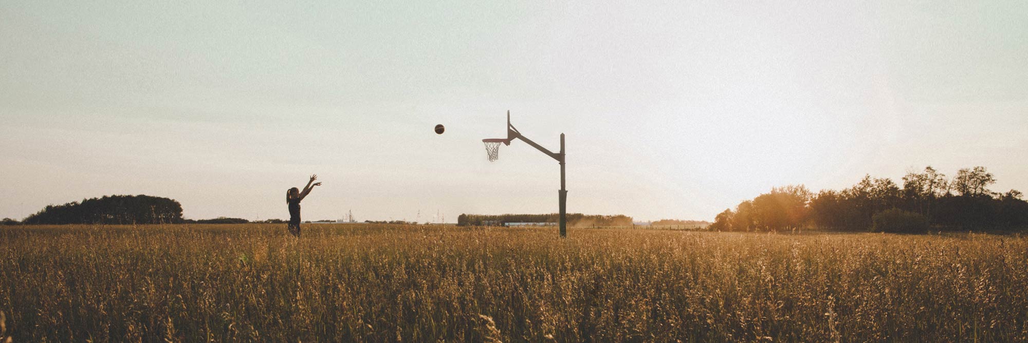 A young woman stands in a rural field and shoots a basketball at a hoop
