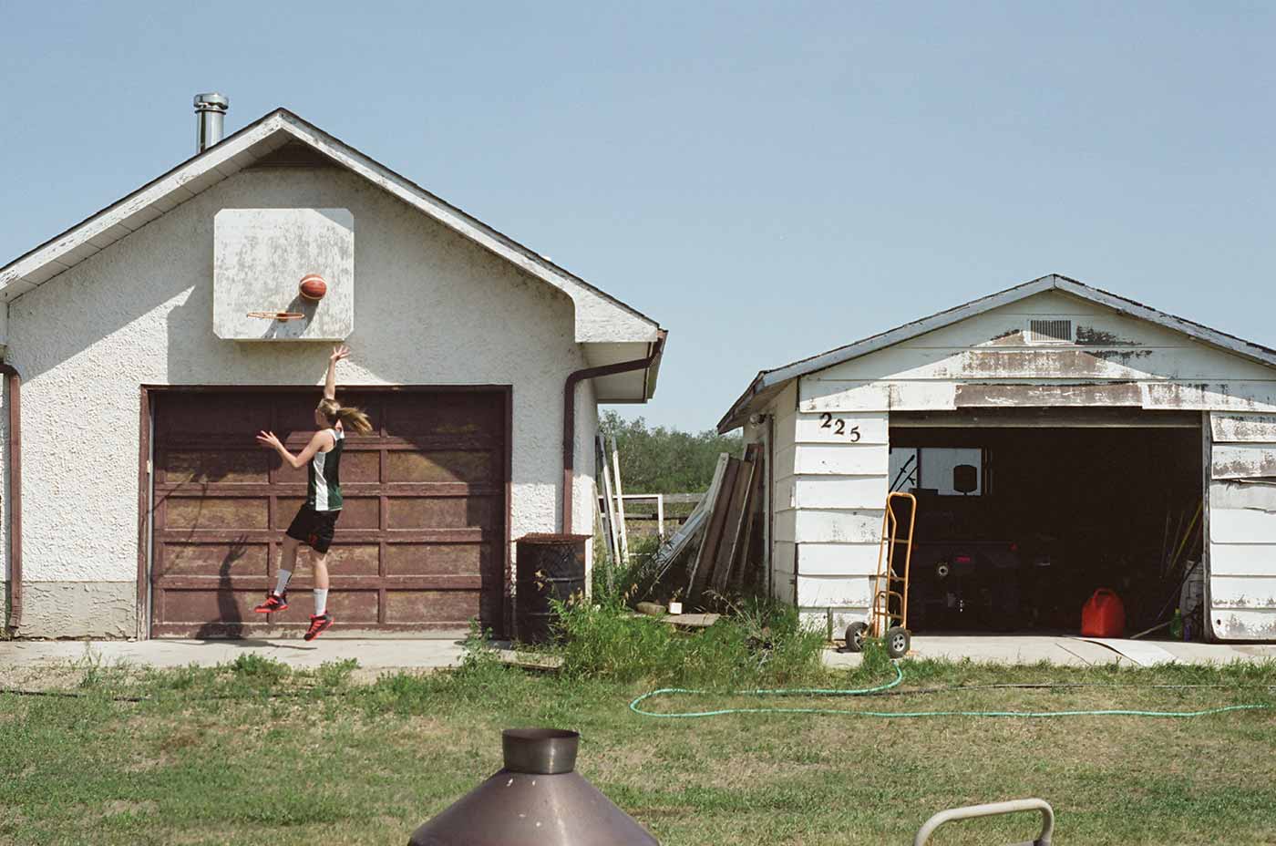 Sydney Tabin shoots a basketball into a net hung above a garage door. The garage and nearby buildings are well weathered and in a rural location.