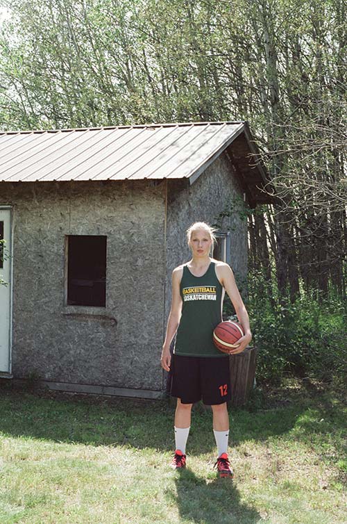 Sydney Tabin poses with a basketball in a Basketball Saskatchewan shirt. She stands in front of a worn down shack.