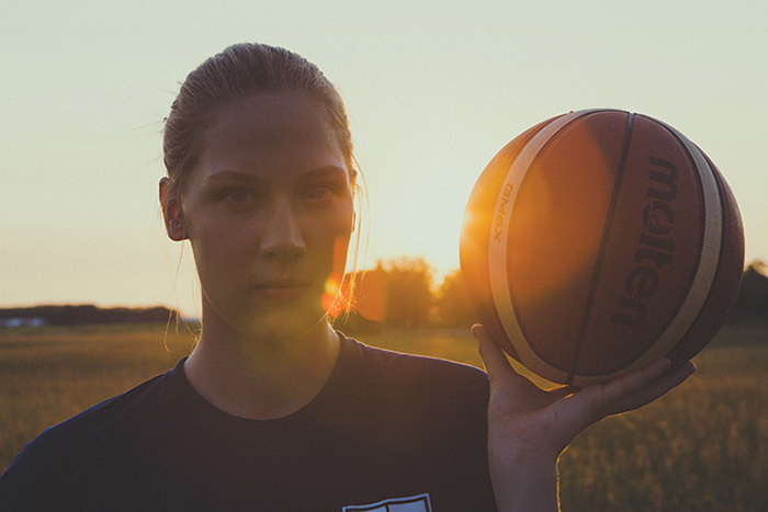 The sun dramatically backlights Sydney Tabin as she stares fiercely at the camera and holds a basketball up by her head.