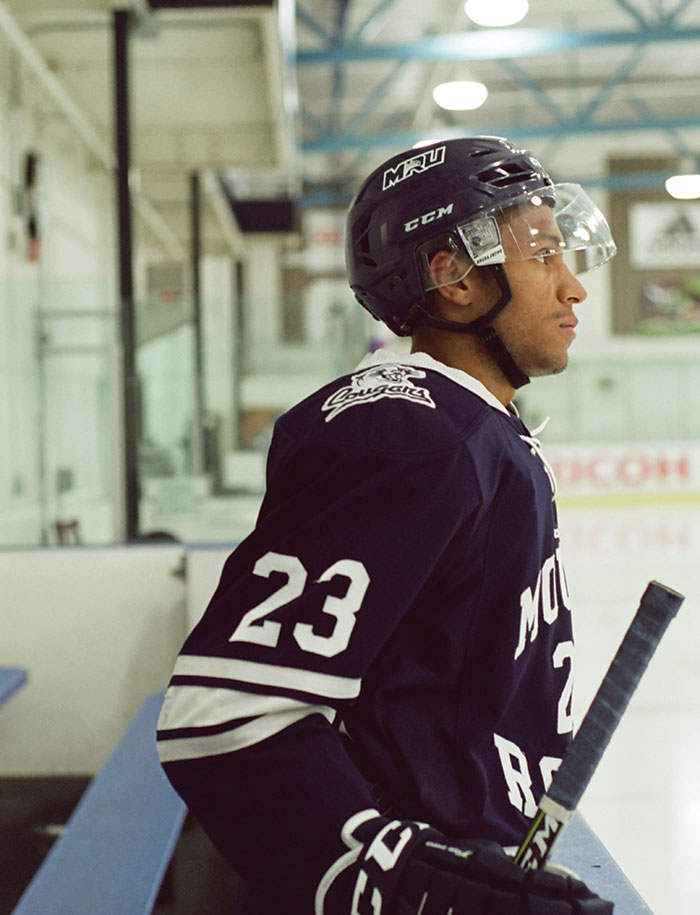 Mount Royal Cougars Men's Hockey player, Jamal Watson, leans against the rink edge waiting to hit the ice.